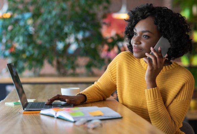 Young black businesswoman having conversation with clients on phone, working at cafe, using laptop, free space