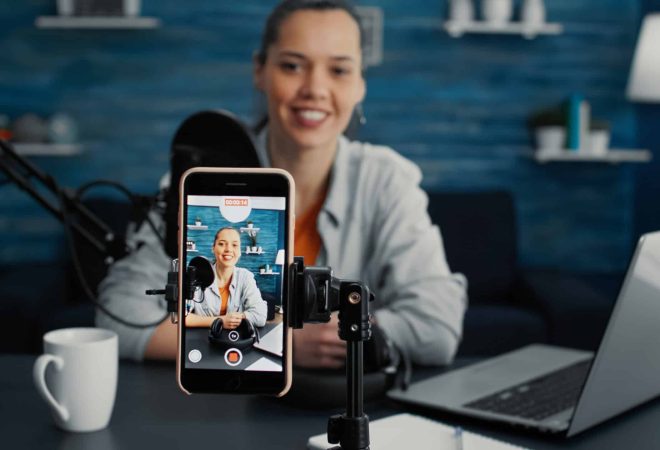 Attractive social media influencer sitting at home studio desk while starting streaming from living room with smartphone. Young content creator recording daily vlog with touchscreen modern device.