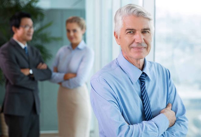 Closeup portrait of content senior business man looking at camera and standing with arms crossed and blurred colleagues in background