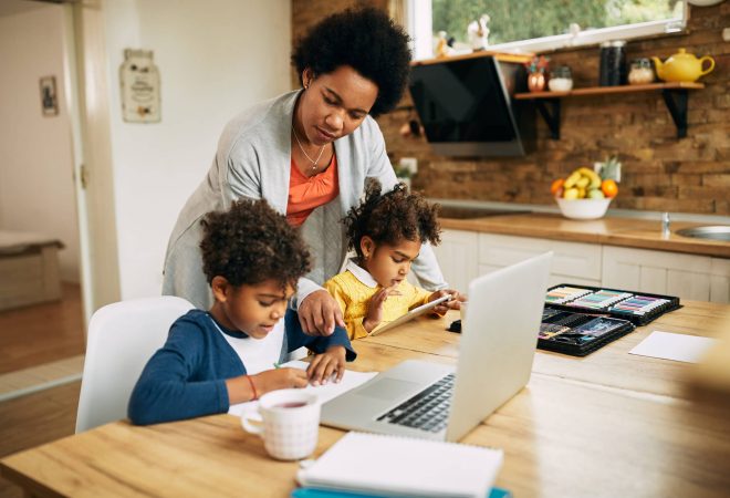 Black single mother helping her kids with learning at home. Small boy is writing on the paper while girl is using touchpad.