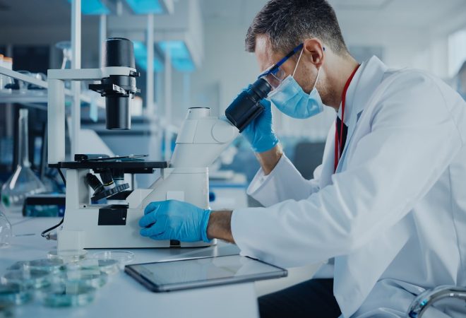 Medical Development Laboratory: Scientist Wearing Face Mask Looking Under Microscope and Using Digital Tablet. Specialists Working on Medicine, Biotechnology Research in Advanced Pharma Lab