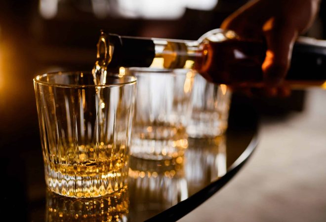 Close up of anonymous man pouring gold whiskey from bottle in glass which standing on wooden table indoors