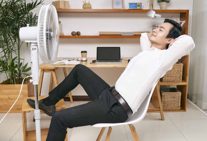 Young Asian office worker resting on chair in front of ventilator and refreshing from heat at office