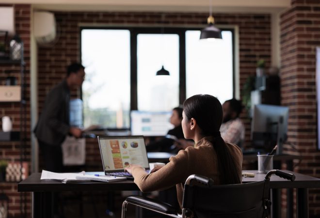 Businesswoman living with disability analyzing report on laptop, working with statistics in disability friendly office. Asian employee suffering from chronic impairment, using wheelchair.