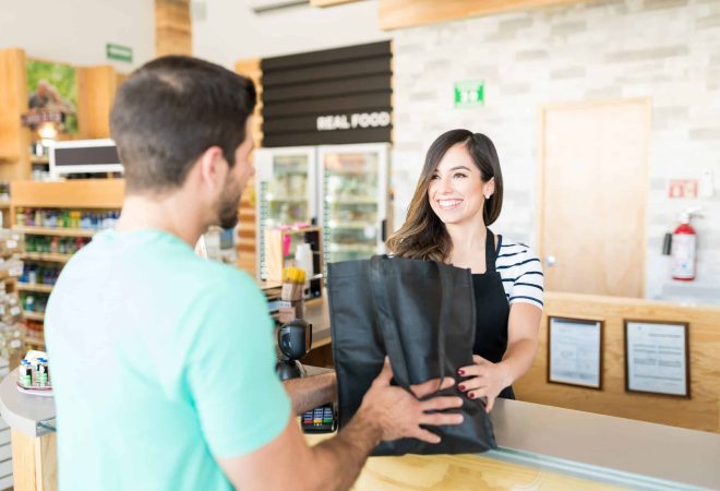 Confident female cashier giving bag to young man at checkout counter in supermarket