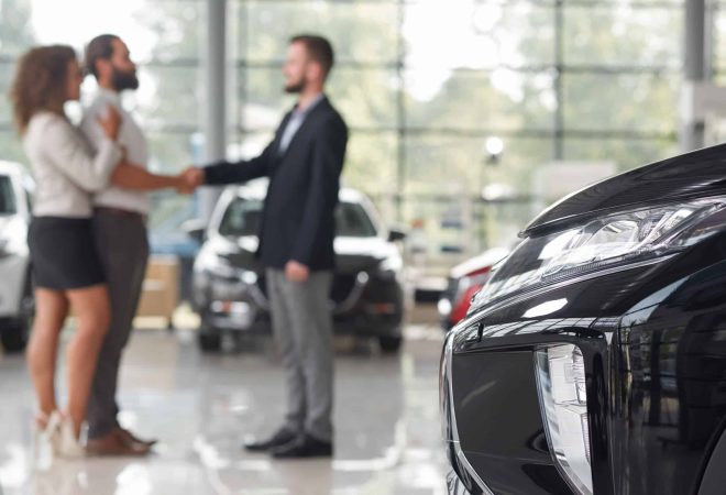 Close up of black car headlight. Couple making deal with car dealer behind. Man and manager of car showroom shaking hands, standing opposite. Purchase of vehicle.