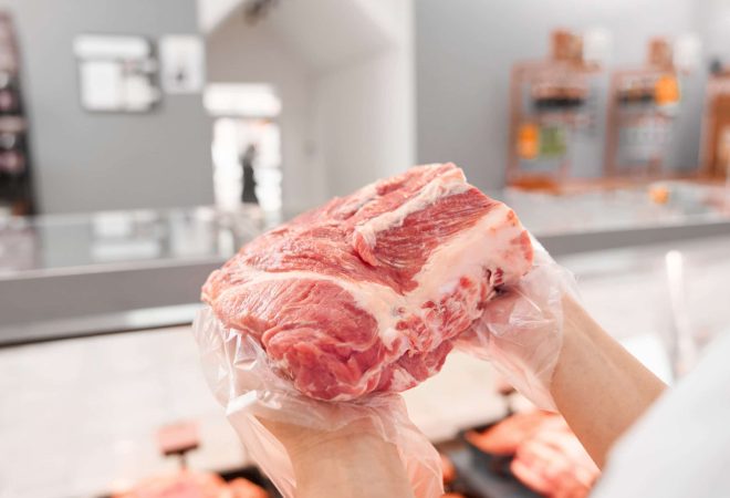 Selective focus of fresh red raw piece of meat in hands of young incognito woman. From above view of girl standing behind counter with big variety of meat and showing pork from refrigerator.