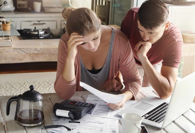 Worried couple reviewing their family finances and calculating expenses, trying to save some money to pay off loan in bank, reviewing bills together, using laptop and calculator at kitchen table