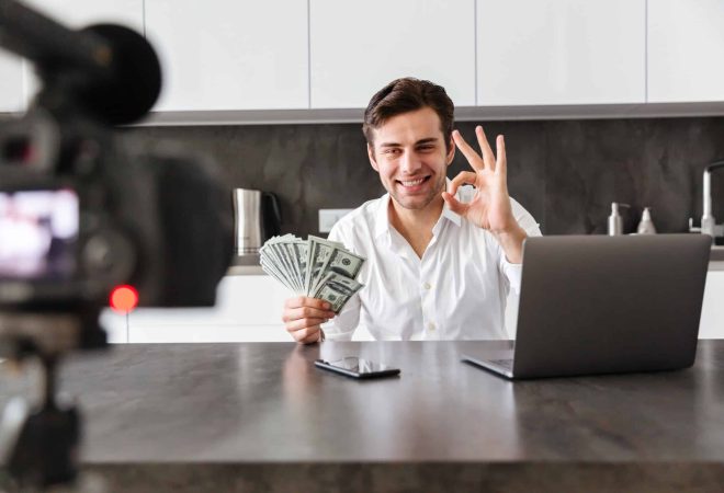 Cheerful young man filming his video blog episode about new tech devices while sitting at the kitchen table with laptop and showing bunch of money banknotes