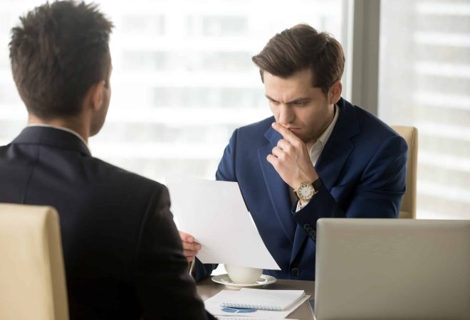 Doubting businessman reading contract document with suspicion when sitting at desk in front of business partner. Entrepreneur unsure in terms of agreement, feeling skeptical because of financial plan