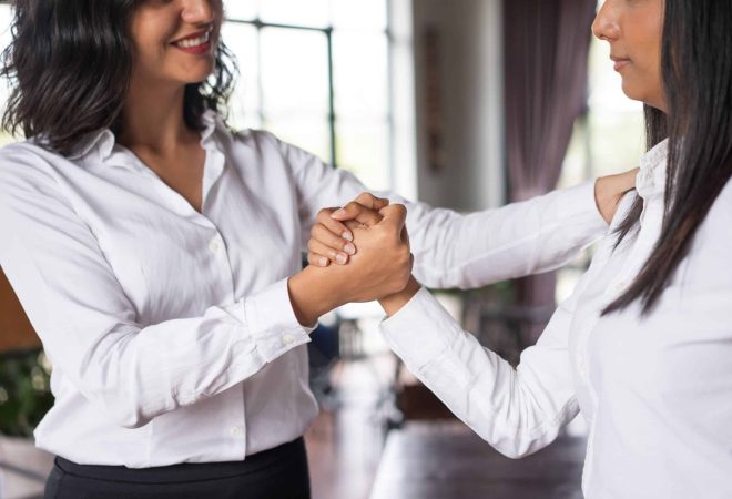 Closeup of smiling business woman supporting colleague in cafe. They are clasping hands and standing with blurred windows in background. Support concept. Side view.