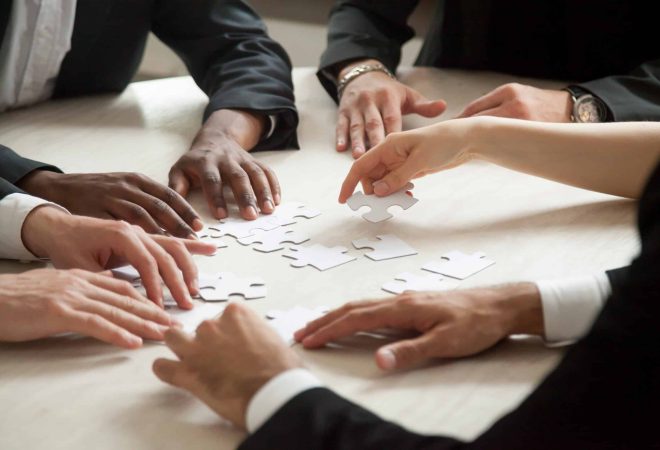 Close up of multiethnic team solving blank puzzle on table. Female hand offering puzzle piece, problem solution concept. Multi-racial executive employees brainstorming about work related problem.