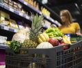 Close up view of shopping cart overloaded with food while in background female person choosing products.