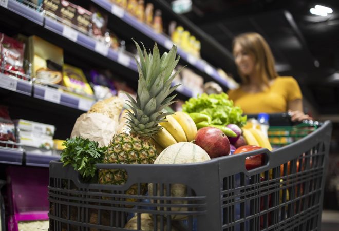 Close up view of shopping cart overloaded with food while in background female person choosing products.