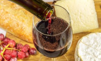 A closeup of red wine poured into a glass at a tasting, with cheese and grapes in the background. Selective focus