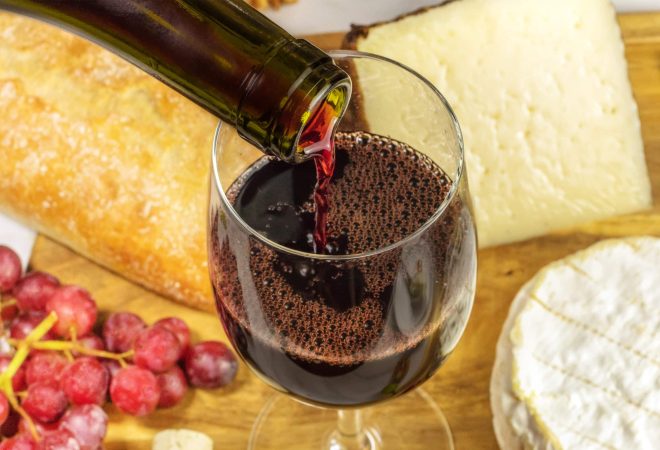 A closeup of red wine poured into a glass at a tasting, with cheese and grapes in the background. Selective focus