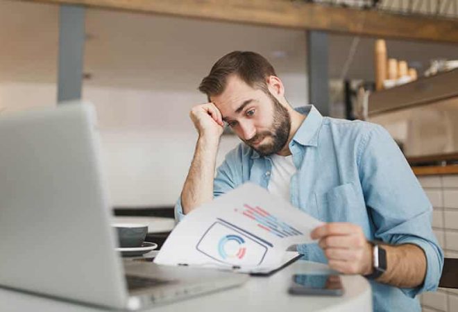 Perplexed puzzled young man sitting alone at table in coffee shop cafe restaurant indoors working or studying on laptop pc computer with papers document. Freelance mobile office business concept