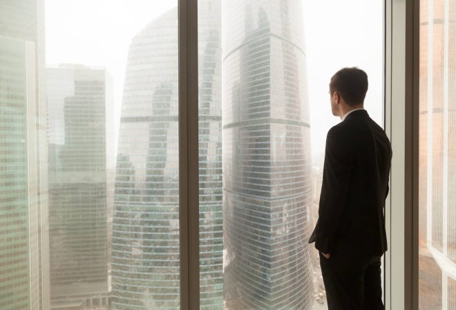 Man in business suit standing with hands in pants pockets looking through window on city landscape with skyscrapers. Businessman thinking about perspectives, dreaming of company success. Back view