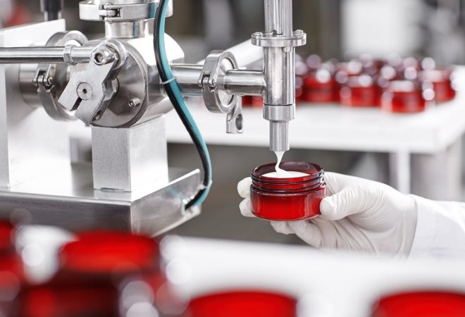 Production and packaging of cosmetics products. Cropped shot of factory worker in gloves and gown holding plastic jar, filling it with white face or body cream. Beauty, skincare and cosmetology