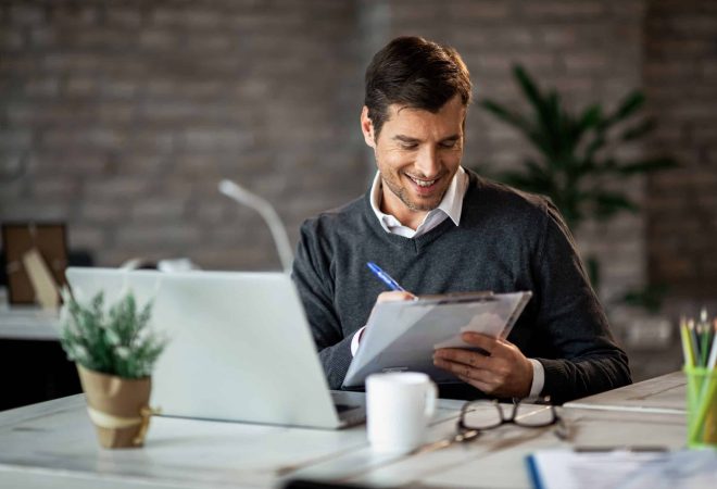 Smiling entrepreneur writing business report while working on laptop in the office.