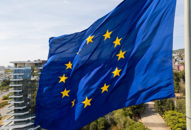 The European Union flag waves in the foreground with a modern cityscape in the background. The dynamic contrast between the flag and urban environment symbolizes unity and progress.