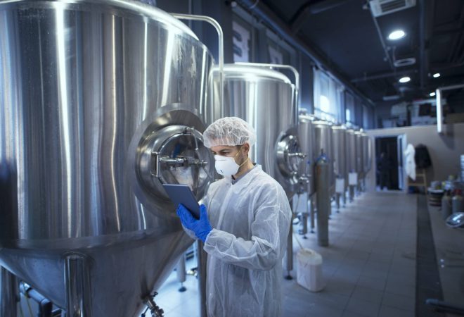 Factory worker technologist in protective uniform with hairnet and mask controlling food production on tablet computer.