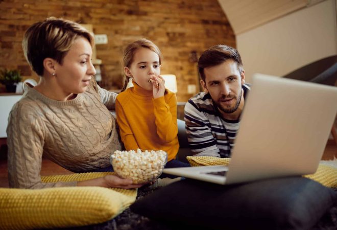 Mid adult parents and their small daughter surfing the net on a computer while eating popcorn at home.