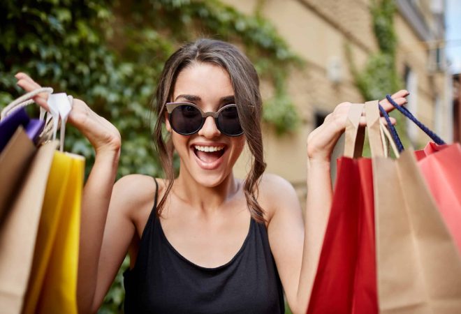 Close up portrait of happy joyful young dark-haired caucasian woman in tab glasses and black shirt looking in camera with opened mouth and happy expression, holding colorful shopping bags in hands. Girl excited with her new clothes