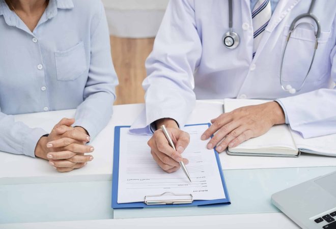 Close-up shot of male doctor wearing white coat filling in medical history while sitting at desk next to female patient