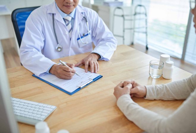 Close-up shot of male doctor filling in medical record, his patient sitting opposite him