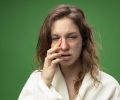 weak young ill girl looking at camera wearing white robe wiping eye with finger isolated on green background