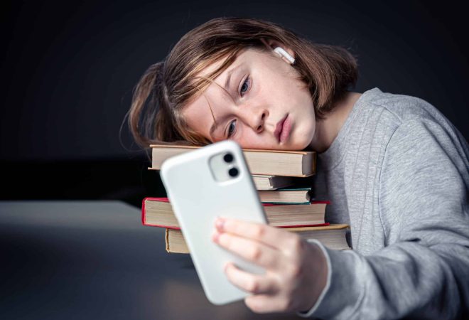 Teenage girl sits with books, but uses a smartphone, boredom from reading, addiction to technology.