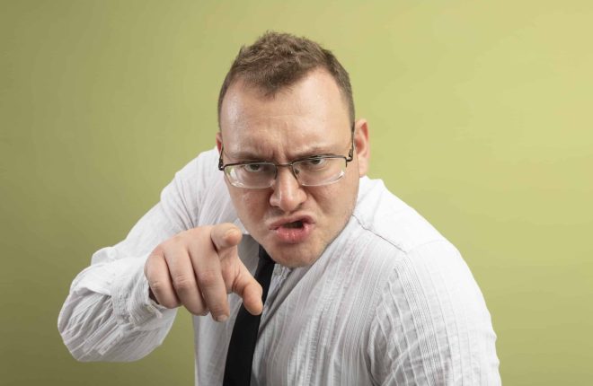 frowning adult slavic man wearing glasses and tie looking and pointing at camera isolated on olive green background