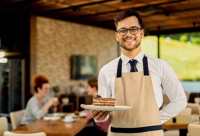 Happy waiter with slice of cake standing in a cafe and looking at camera. There are people in the background.