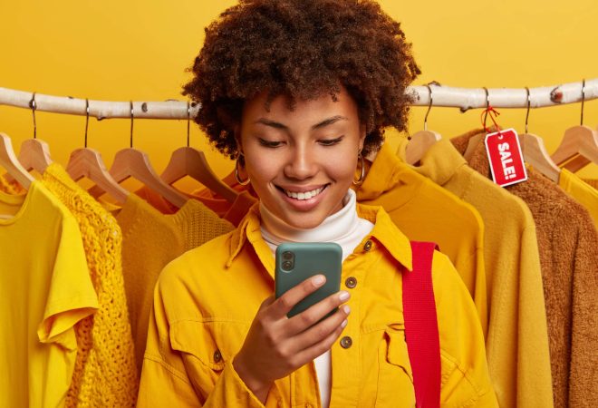 Glad online merchant focused in smartphone device, enjoys biggest shopping day, rejoices marked down prices, stands against yellow clothing on racks, celebrates importnat ecommerce holiday in autumn