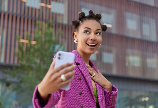 Happy dreamy woman with funny hairstyle makes photo of herself wears fashionable pink jacket looks somewhere positively poses outdoors against blurred background. Technology and urban lifestyle