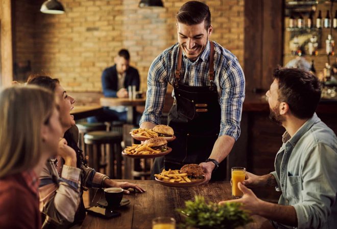 Group of happy friends having fun while waiter is serving them food in a pub. Focus is on waiter.
