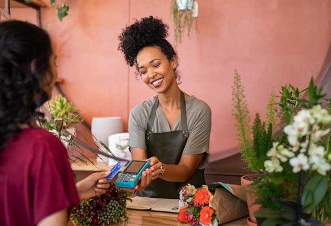 Smiling and friendly florist holding card reader machine at counter with customer paying with credit card. Young african american florist shop assistant holding payment machine while buyer purchase a bunch flower. Woman using bank credit card to make payment by NFC machine.