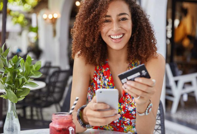 Internet banking and ecommerce concept. Happy young smiling female with Afro hairstyle, uses modern cell phone and credit card for online shopping, enjoys fresh fruit smoothie in terrace bar.