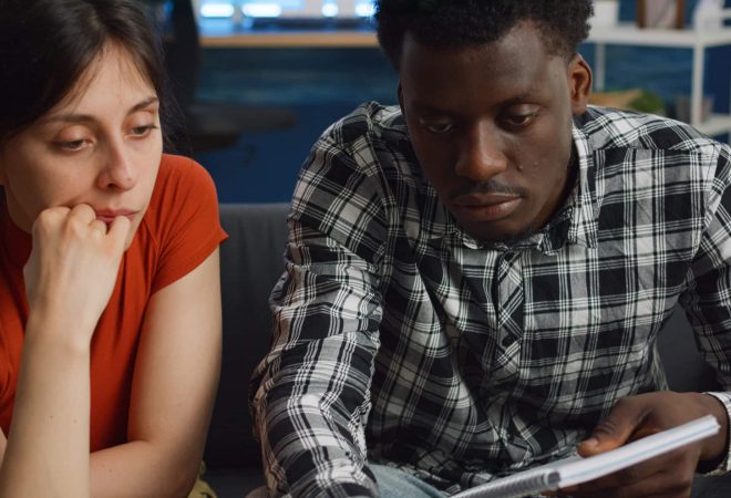 Interracial people paying taxes and bills checking budget on laptop at home. Mixed race couple calculating tax money writing in notebook for financial planning on living room couch