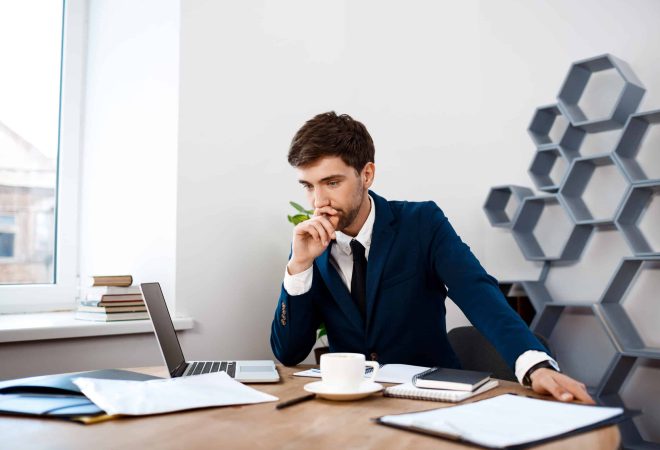 Young successful businessman in suit sitting at workplace, thinking, office background.