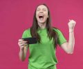 young beautiful girl wearing green t-shirt holding smartphone crazy happy raising clenched fist smiling cheerfully rejoicing her success standing over isolated pink background