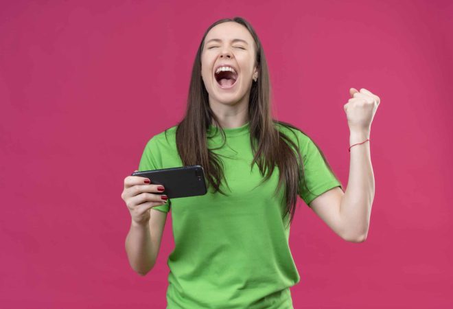 young beautiful girl wearing green t-shirt holding smartphone crazy happy raising clenched fist smiling cheerfully rejoicing her success standing over isolated pink background