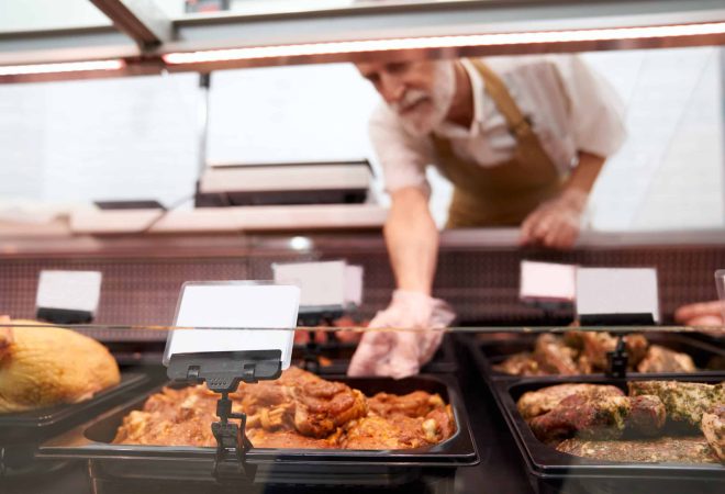 Close up of raw meat pieces in refrigerator with price tags ready for sale in meat department. Senior male butcher putting out of glass counter plate with sliced fresh steaks. Concept of food.