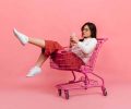 Stylish teen girl in culottes and white sweater sits in supermarket trolley. Model in glasses sends kiss and makes selfie on pink