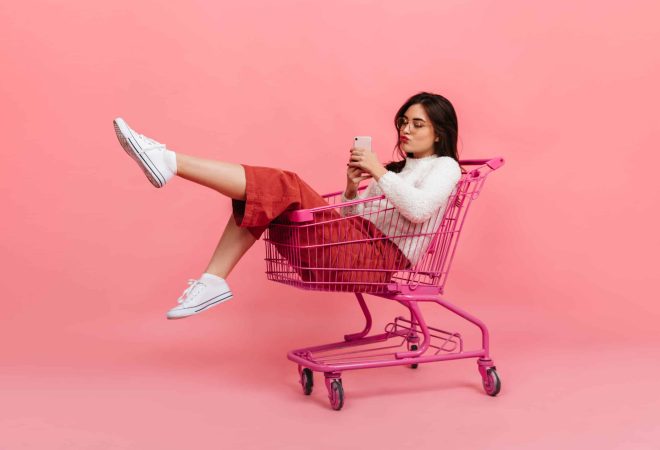 Stylish teen girl in culottes and white sweater sits in supermarket trolley. Model in glasses sends kiss and makes selfie on pink