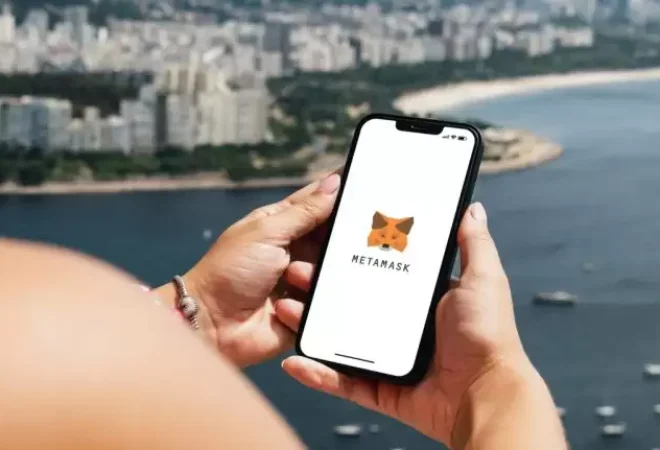 mulher-segura-celular-com-logo-da-metamask-com-paisagem-de-praia-ao-fundo-750x431