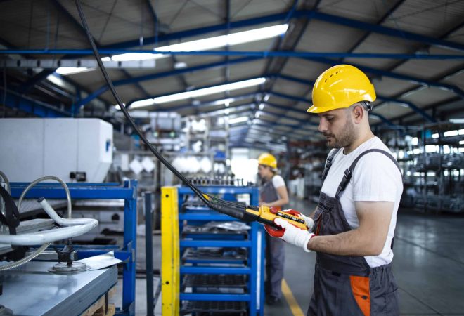 Factory worker wearing uniform and hardhat operating industrial machine with push button joystick in production hall.