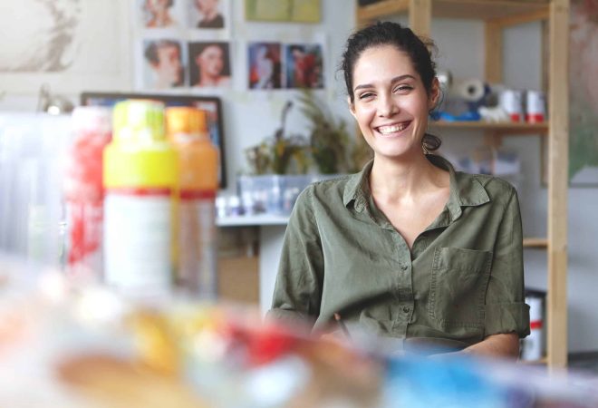 Picture of charming charismatic young female artist wearing khaki shirt grinning broadly feeling happy about her job and creating process, sitting at workshop, surrounded with painting accessories