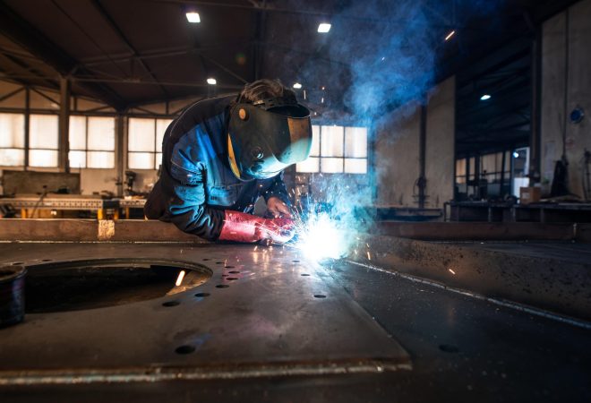 Professional industrial welder welding metal parts in metalworking factory.